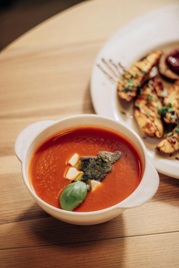 A warm tomato soup bowl garnished with basil and cheese cubes beside grilled bread.