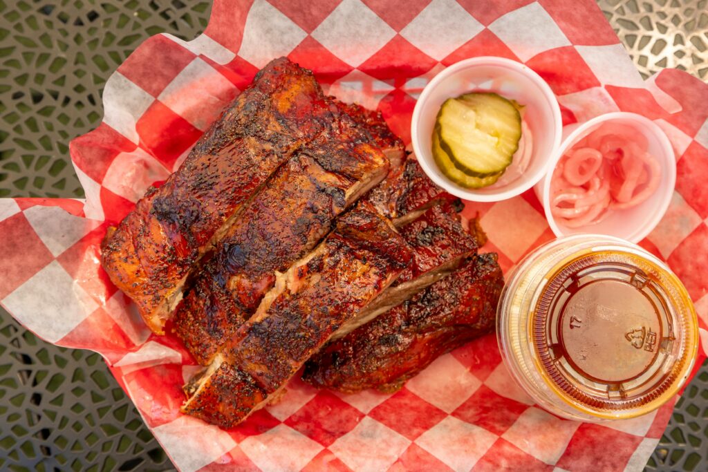 Close-up of grilled barbecue ribs with pickles, onions, and sauce on picnic table.