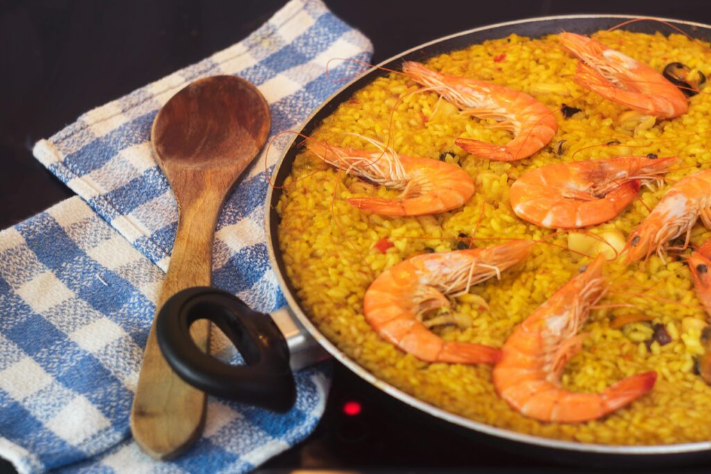 Close-up of a seafood paella with prawns in a pan, a wooden spoon, and blue towel.