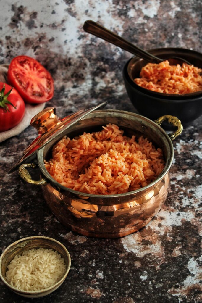 Appetizing tomato rice served in a copper pot with sliced tomatoes and raw grains on a rustic surface.